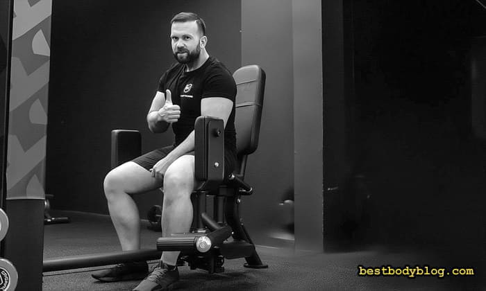 Мускулистий чоловік сидить у тренажері для розведення ніг і показує великий палець вгору, висловлюючи схвалення